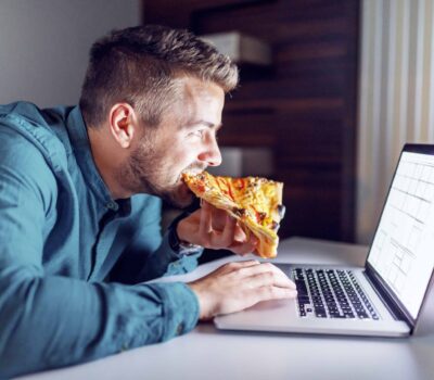 Side view of Caucasian hardworking architect eating pizza and using laptop while sitting in office late at night.
